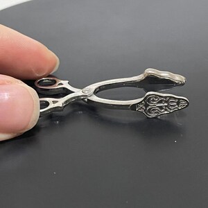 May include: Silver-toned metal tongs with an ornate design. The tongs are closed and have a decorative handle.