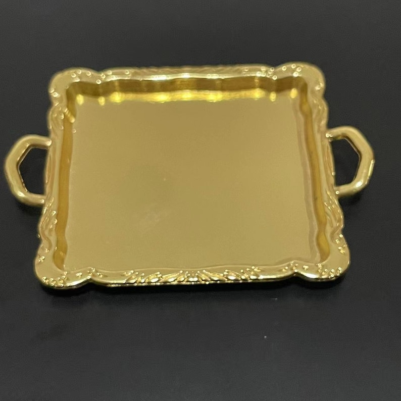 May include: A gold-colored metal serving tray with two handles. The tray has a decorative border and a smooth, reflective surface.
