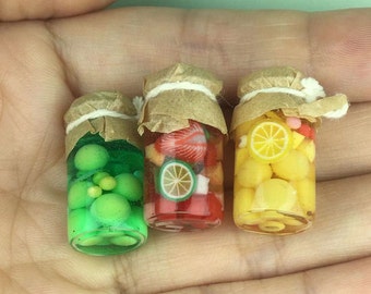 Può includere: Tre vasetti di vetro in miniatura con coperchi di carta marrone e legature di spago, riempiti di fette di frutta colorate. I vasetti contengono uva verde, fragole rosse e ananas, e limoni gialli.