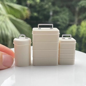 Puede incluir: Tres contenedores de almacenamiento de metal blanco con asas plateadas. Los contenedores son de diferentes tamaños y están apilados uno encima del otro.