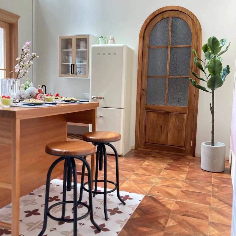 May include: A white refrigerator with a wood-toned cabinet above it. There are three bar stools with black metal frames and wood seats in front of a wooden kitchen island. A potted plant is in the corner of the room, and a wooden arched door with glass panels is in the background.