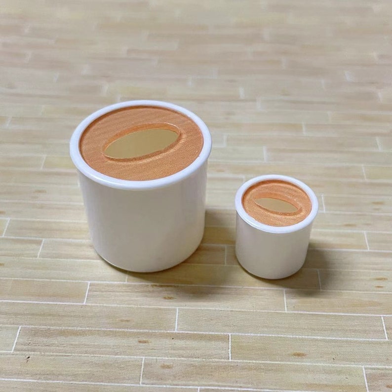May include: Two white ceramic containers with orange lids. The containers have a round shape and a hole in the center of the lid. The larger container is about 3 inches tall and the smaller container is about 2 inches tall.