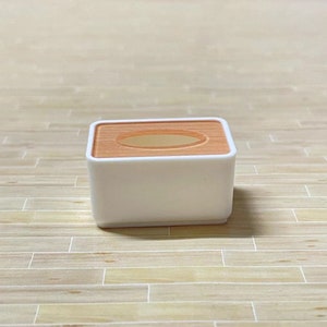 May include: A white tissue box with a light brown top and a rectangular opening. The box is sitting on a light brown wooden surface.
