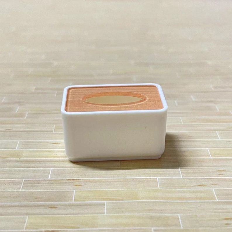 May include: A white tissue box with a light brown top and a rectangular opening. The box is sitting on a light brown wooden surface.