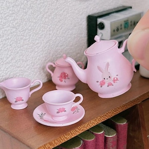 May include: A pink tea set with a teapot, creamer, sugar bowl, and teacup and saucer. The teapot has a bunny design and the other pieces have floral designs.