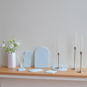 May include: A set of six light blue decorative trays with various geometric patterns. The trays are made of a textured material and are arranged on a wooden shelf. Three silver candle holders with white candles are also on the shelf.
