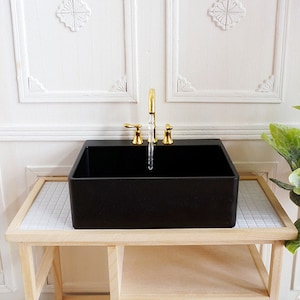 May include: A miniature black rectangular sink with a gold faucet. The sink is set on a light wood table with a white and gray tiled surface. The background features white paneling.