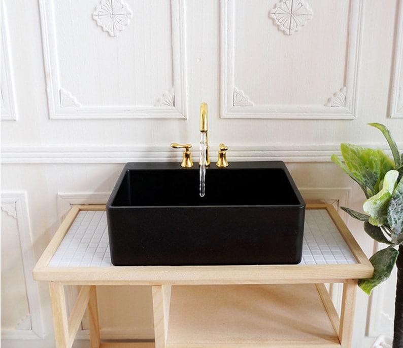May include: A miniature black rectangular sink with a gold faucet. The sink is set on a light wood table with a white and gray tiled surface. The background features white paneling.