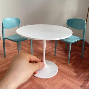 May include: A miniature white round table with a single stem base. Two light blue chairs are in the background. The table is approximately 30 cm in diameter, suitable for a dollhouse or miniature setting.
