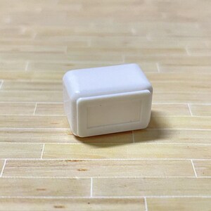 May include: A small white plastic box with a rectangular opening on the front. The box is sitting on a light brown wooden surface.