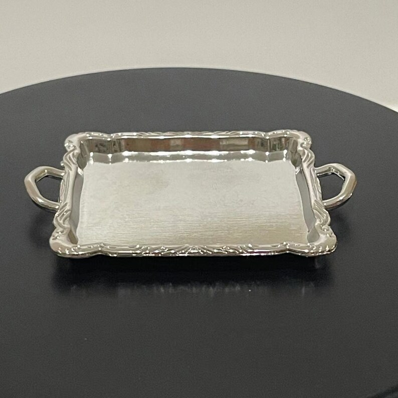 May include: A silver serving tray with ornate edges and two handles. The tray is empty and has a reflective surface.