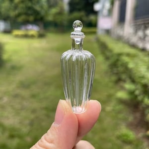 Puede incluir: Una botella de vidrio transparente con un diseño acanalado y un tapón redondo. La botella se sostiene en una mano.