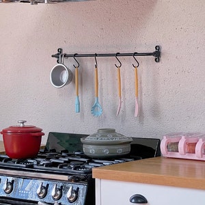 May include: A black metal rack with four hanging utensils and a white ceramic mug. The rack is mounted on a white wall above a black and silver stovetop. A red pot and a gray ceramic bowl are on the stovetop.