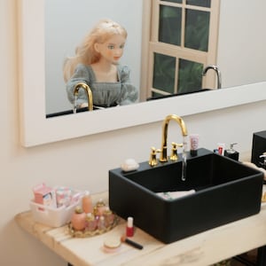 May include: A miniature bathroom scene featuring a black sink with a gold faucet and a mirror reflecting a doll. The sink has a soap dish, cosmetics, and a tube of lotion. A white basket holds more toiletries, and a tray displays perfume bottles.
