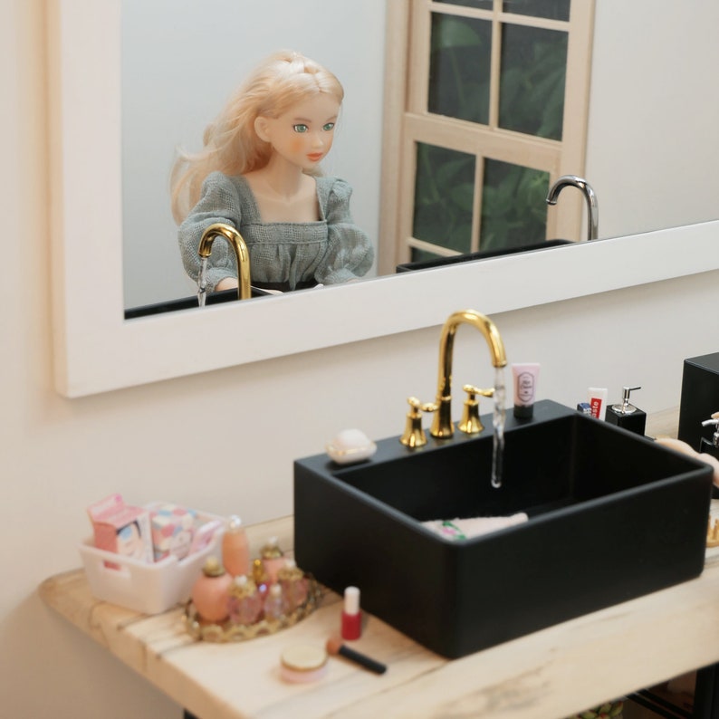 May include: A miniature bathroom scene featuring a black sink with a gold faucet and a mirror reflecting a doll. The sink has a soap dish, cosmetics, and a tube of lotion. A white basket holds more toiletries, and a tray displays perfume bottles.