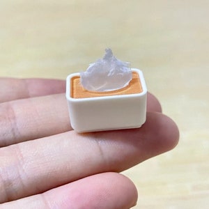 May include: A small white square container with a light orange rim, holding a white, translucent, waxy substance.