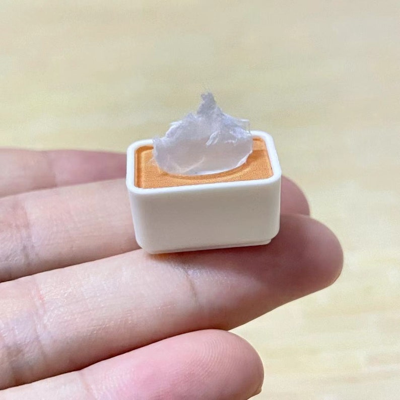 May include: A small white square container with a light orange rim, holding a white, translucent, waxy substance.