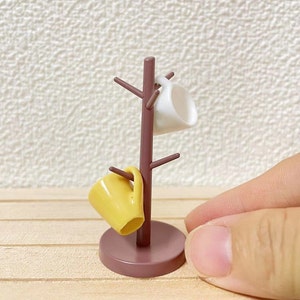 Może przedstawiać: Miniaturowy drewniany stojak na kubki z brązową podstawą i brązowym drewnianym słupkiem. Na stojaku wiszą dwa kubki, jeden jest żółty, a drugi biały.
