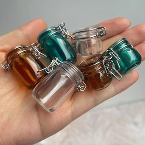 Puede incluir: Conjunto de seis pequeños frascos de vidrio con tapas de metal. Los frascos son transparentes, marrones y verde azulado. Las tapas tienen un cierre con bisagras.