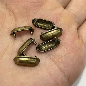 May include: A collection of antique brass-toned metal drawer pulls. Each pull is rectangular with rounded edges and two small posts for attachment. The pulls have a vintage, aged appearance, suggesting they are for restoration or craft projects.