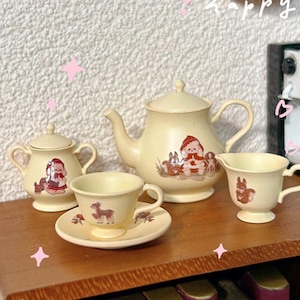 May include: A miniature tea set with a cream-colored teapot, sugar bowl, creamer, and teacup and saucer. The set features a whimsical design with illustrations of animals and a girl in a red hooded cloak.