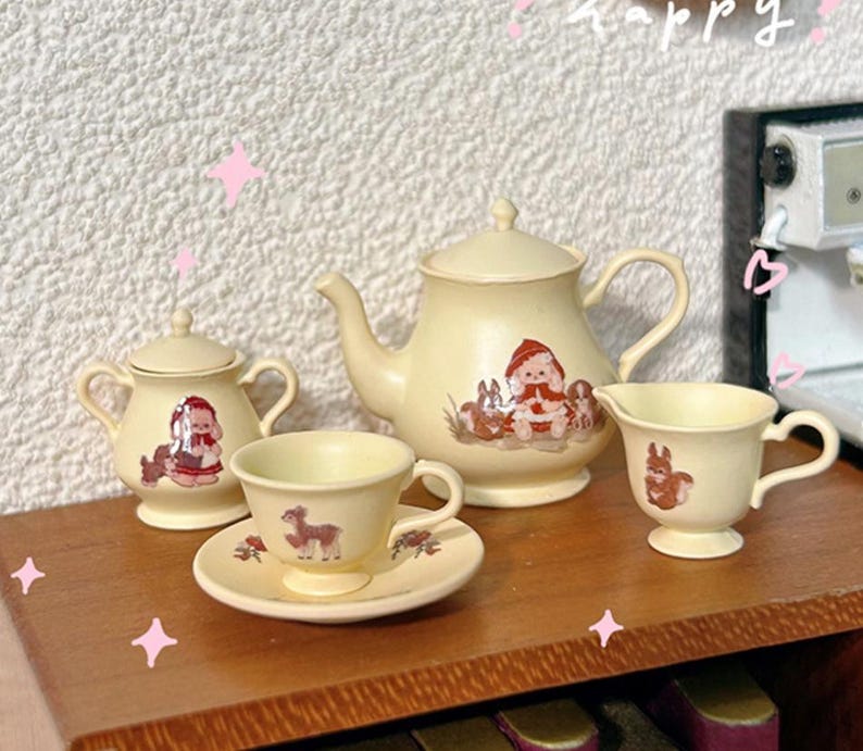 May include: A miniature tea set with a cream-colored teapot, sugar bowl, creamer, and teacup and saucer. The set features a whimsical design with illustrations of animals and a girl in a red hooded cloak.