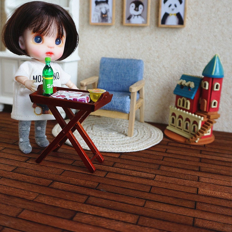May include: A doll with blue eyes and brown hair is holding a tray with a bottle of green soda, a box of snacks, and a small bowl. The tray is on a wooden folding table. The doll is wearing a white shirt with the word "MO" on it and grey tights. The doll is standing on a wooden floor.