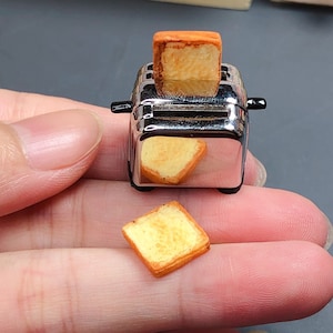 Könnte beinhalten: Ein kleiner silberner Toaster im Miniaturformat mit einer Scheibe Brot im Schlitz und einer weiteren Scheibe Brot daneben.