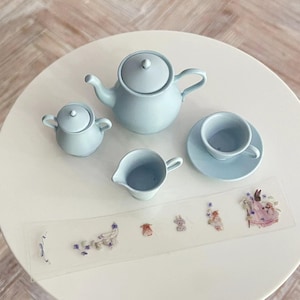 May include: A light blue tea set with a teapot, creamer, sugar bowl, and saucer with a teacup. The set is on a white table with a sheet of paper with small floral stickers.