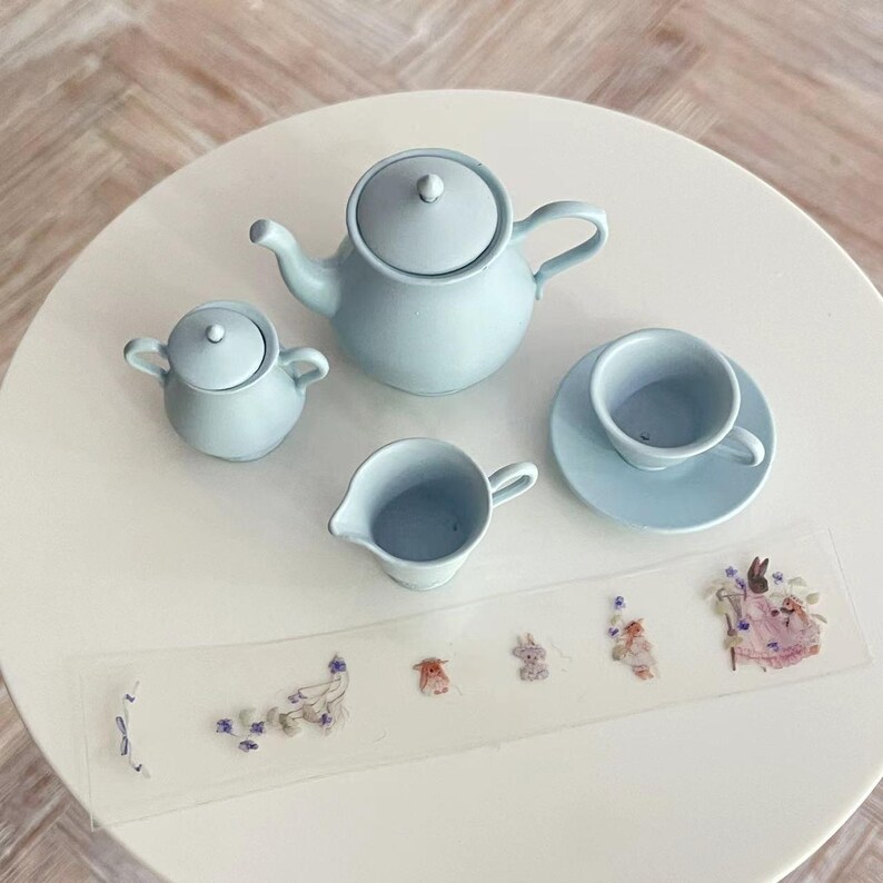 May include: A light blue tea set with a teapot, creamer, sugar bowl, and saucer with a teacup. The set is on a white table with a sheet of paper with small floral stickers.