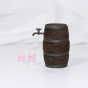 Pode incluir: Um barril de madeira marrom com faixas de metal e uma pequena torneira de metal. Dois copos de vidro transparentes cheios de l&iacute;quido rosa est&atilde;o na frente do barril.