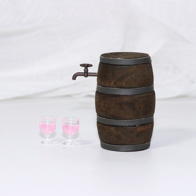 Pode incluir: Um barril de madeira marrom com faixas de metal e uma pequena torneira de metal. Dois copos de vidro transparentes cheios de l&iacute;quido rosa est&atilde;o na frente do barril.