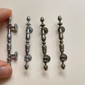 May include: Three ornate metal cabinet door pulls in silver, brass, and bronze finishes. Each pull features a decorative, scroll-like design with a rounded top.