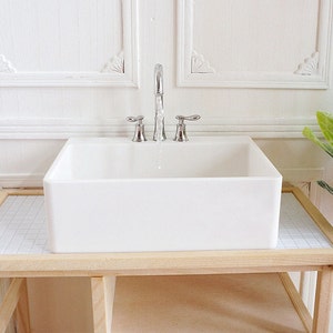May include: A miniature white porcelain sink with a chrome faucet and two handles. The sink is rectangular and sits on a light wood stand with a tiled surface. The background is a white wall with decorative molding.