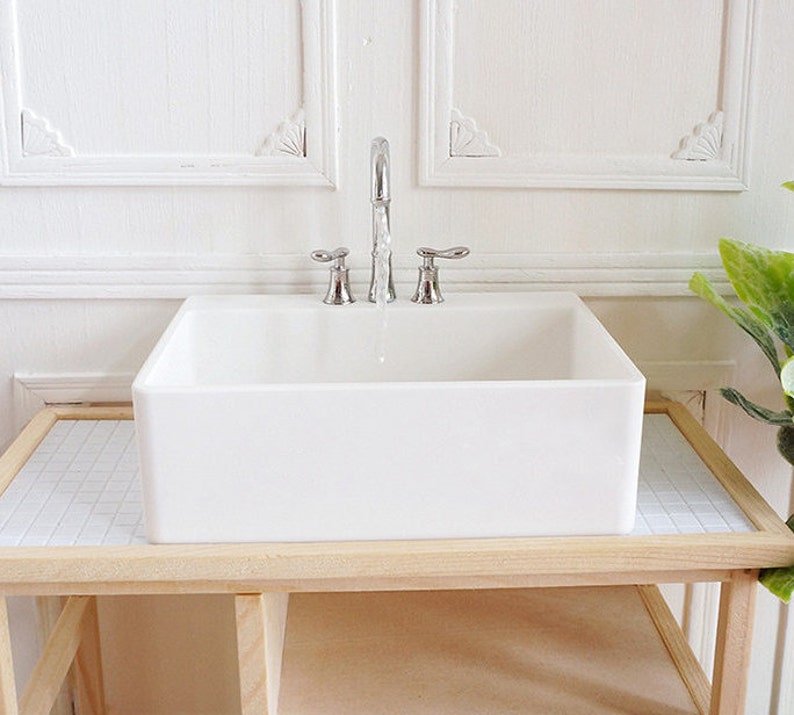 May include: A miniature white porcelain sink with a chrome faucet and two handles. The sink is rectangular and sits on a light wood stand with a tiled surface. The background is a white wall with decorative molding.