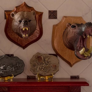 May include: Miniature bear and hippo trophy heads mounted on wooden plaques. Two fossil replicas are displayed below. The bear has an open mouth with visible teeth. The hippo has an open mouth with visible teeth. The fossils are on gold stands.
