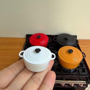 May include: A collection of four miniature round casserole dishes in white, red, black, and orange, each featuring a matching lid with a black knob and two side handles. These small pots are arranged on a black miniature stove with a grate surface.