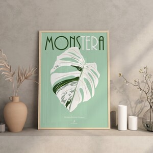 May include: A framed poster with a green background and the word "Monstera" in a stylized font. A white and green Monstera Deliciosa plant leaf is featured in the center of the poster.