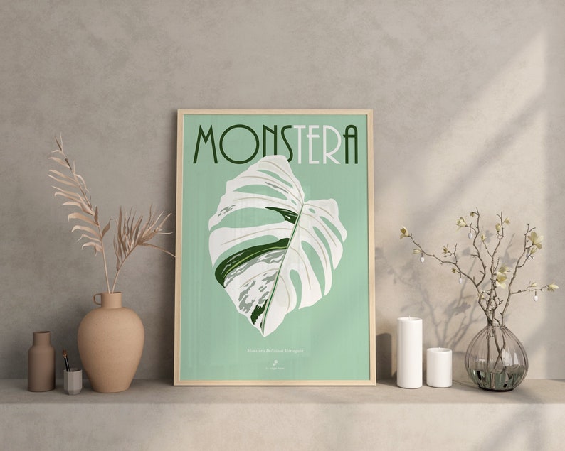 May include: A framed poster with a green background and the word "Monstera" in a stylized font. A white and green Monstera Deliciosa plant leaf is featured in the center of the poster.
