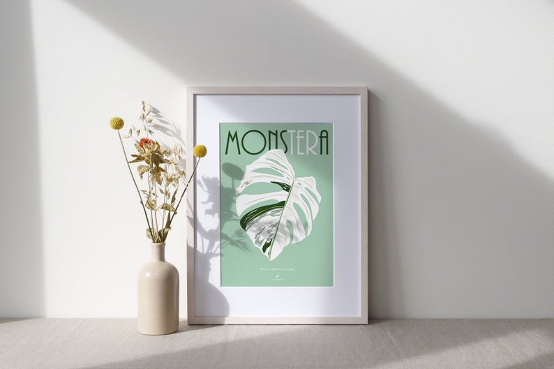 May include: A framed print with a white background and a green border. The print features a white Monstera leaf with green veins against a light green background. The word "Monstera" is written in green letters at the top of the print.