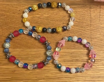 May include: Three beaded bracelets with different colored beads and metal accents. The first bracelet has white, yellow, blue, and black beads. The second bracelet has pink, white, blue, and purple beads. The third bracelet has pink, white, blue, and silver beads.