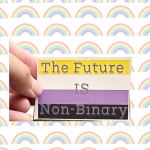 May include: A rectangular sticker with the text "The Future IS Non-Binary" in a non-binary pride flag design. The flag features yellow, white, purple, and black horizontal stripes. The sticker is held against a background of repeating rainbow arcs.