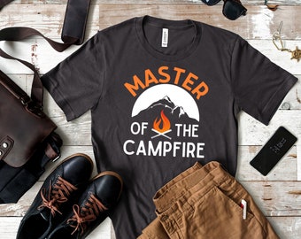 May include: A dark grey t-shirt with the text "Master of the Campfire" in white. The text is surrounded by a white circle with a mountain range and a campfire inside.