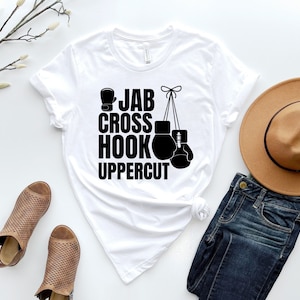 May include: White t-shirt with black text that reads "Jab Cross Hook Uppercut" and a graphic of boxing gloves hanging from a string.