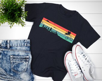 May include: A dark blue t-shirt with a retro sunset graphic featuring a silhouette of a cat riding a bicycle. The graphic is in shades of orange, yellow, green, and blue.