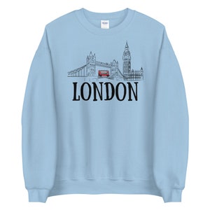 May include: A light blue sweatshirt with a black and white illustration of the London skyline, including Big Ben, Tower Bridge, and a red double-decker bus. The text "LONDON" is printed in black below the illustration.
