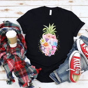 May include: Black t-shirt with a floral pineapple design. The pineapple is made of colorful flowers and is surrounded by a gold glitter effect. The shirt is laid out on a white wooden surface with a plaid scarf, a coffee cup, and a pair of red converse shoes.