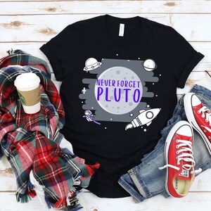 May include: Black t-shirt with a white and gray graphic of a moon with the text "Never Forget Pluto" in purple letters. The graphic also includes a rocket, a spaceship, and stars.