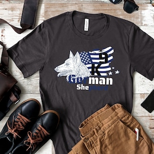 Może przedstawiać: Szary t-shirt z białym nadrukiem głowy owczarka niemieckiego z niebiesko-białą flagą amerykańską z tyłu. Pod głową psa znajduje się napis "911 K9 Man She Protects".