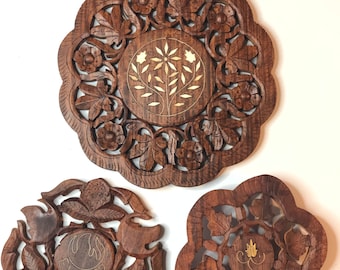 May include: Three intricately carved wooden coasters with floral designs. The coasters are round and have a raised edge. The center of each coaster features a different design, including a tree, a flower, and a crown. The coasters are made of dark brown wood and have a natural finish.